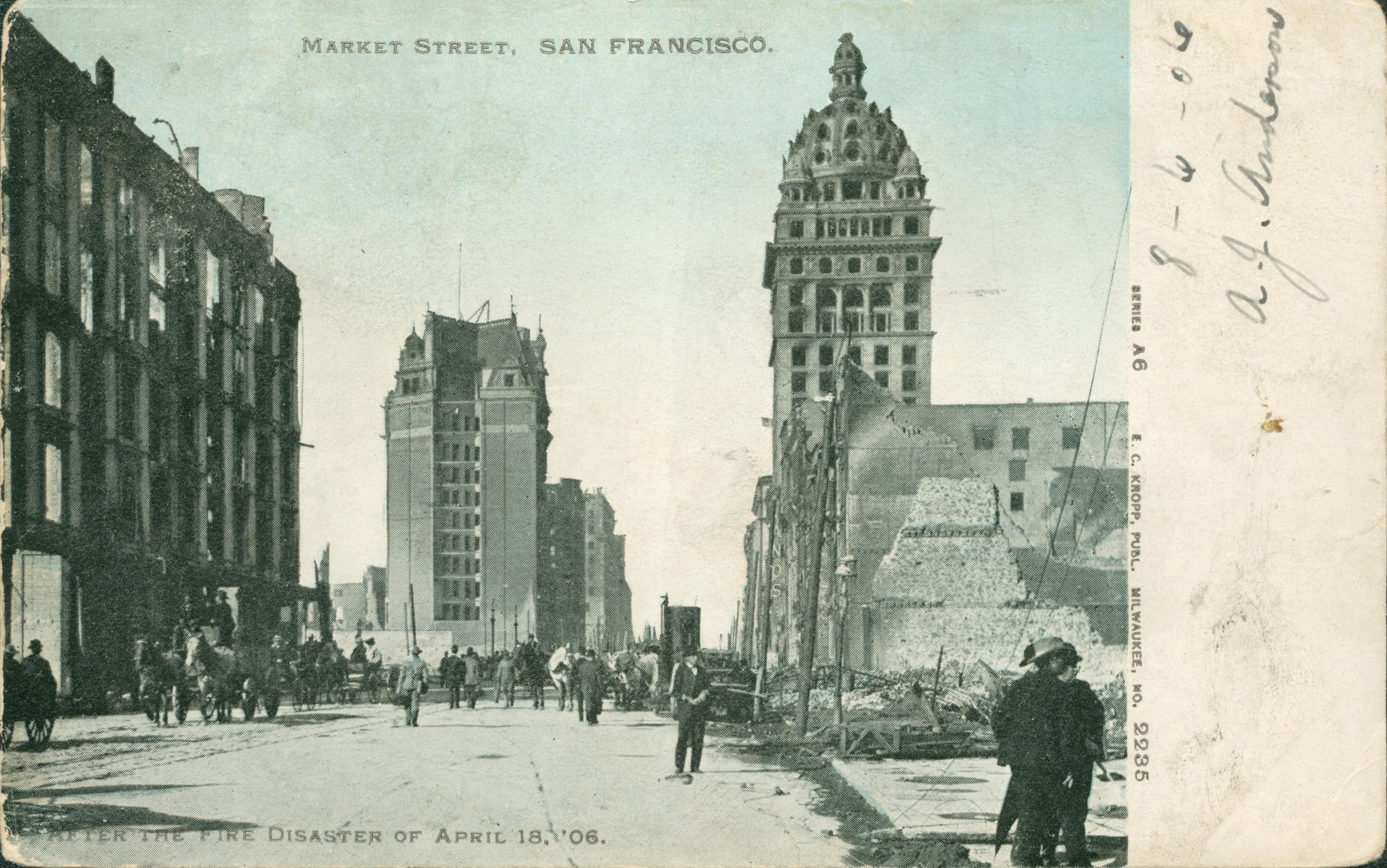 Shows people walking down Market Street with ruined buildings along both sides of the street.