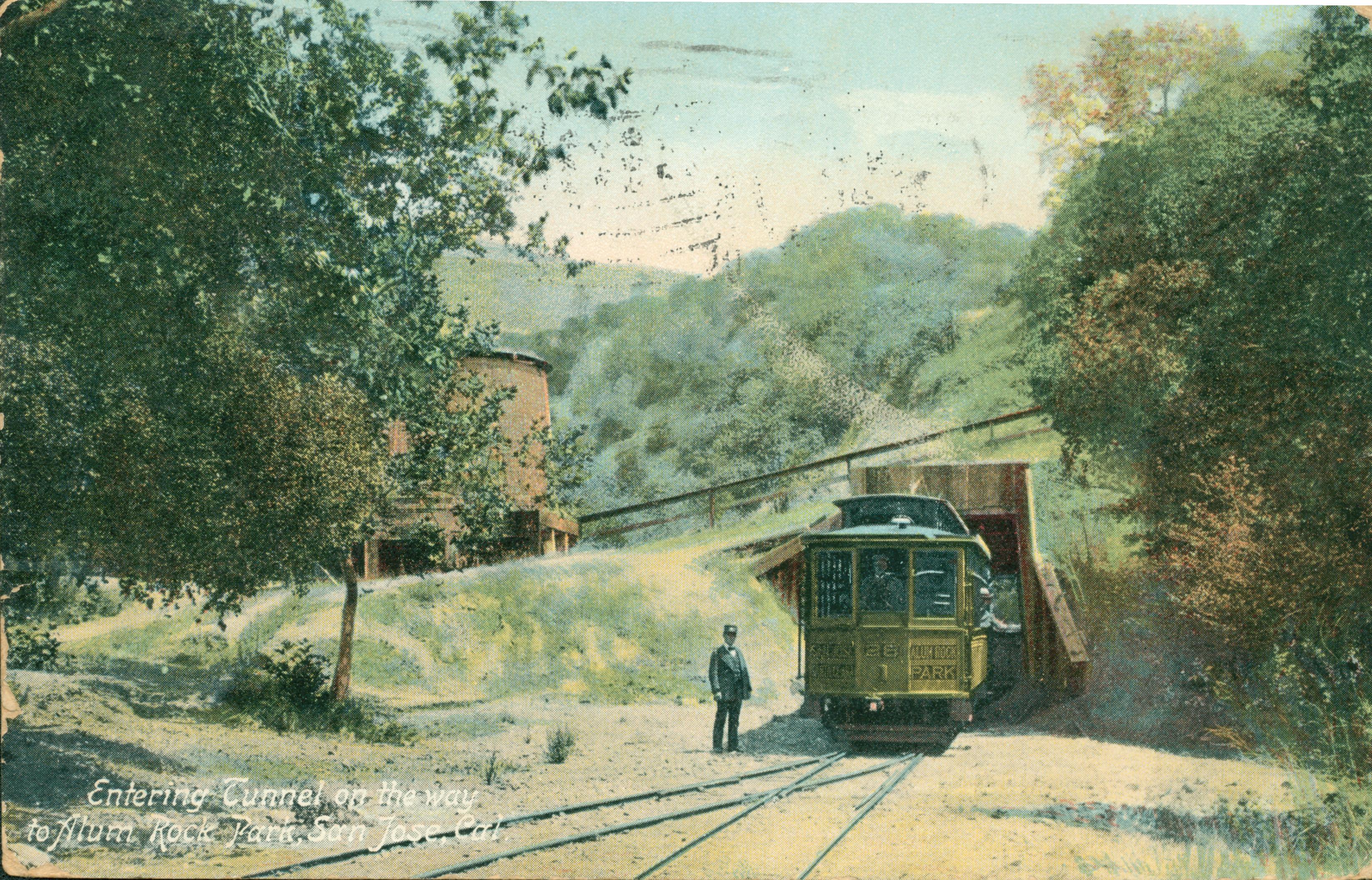 Color view of train and conductor heading into a tunnel on the way to Alum Rock Park, trees, hillside and water tower in the background
