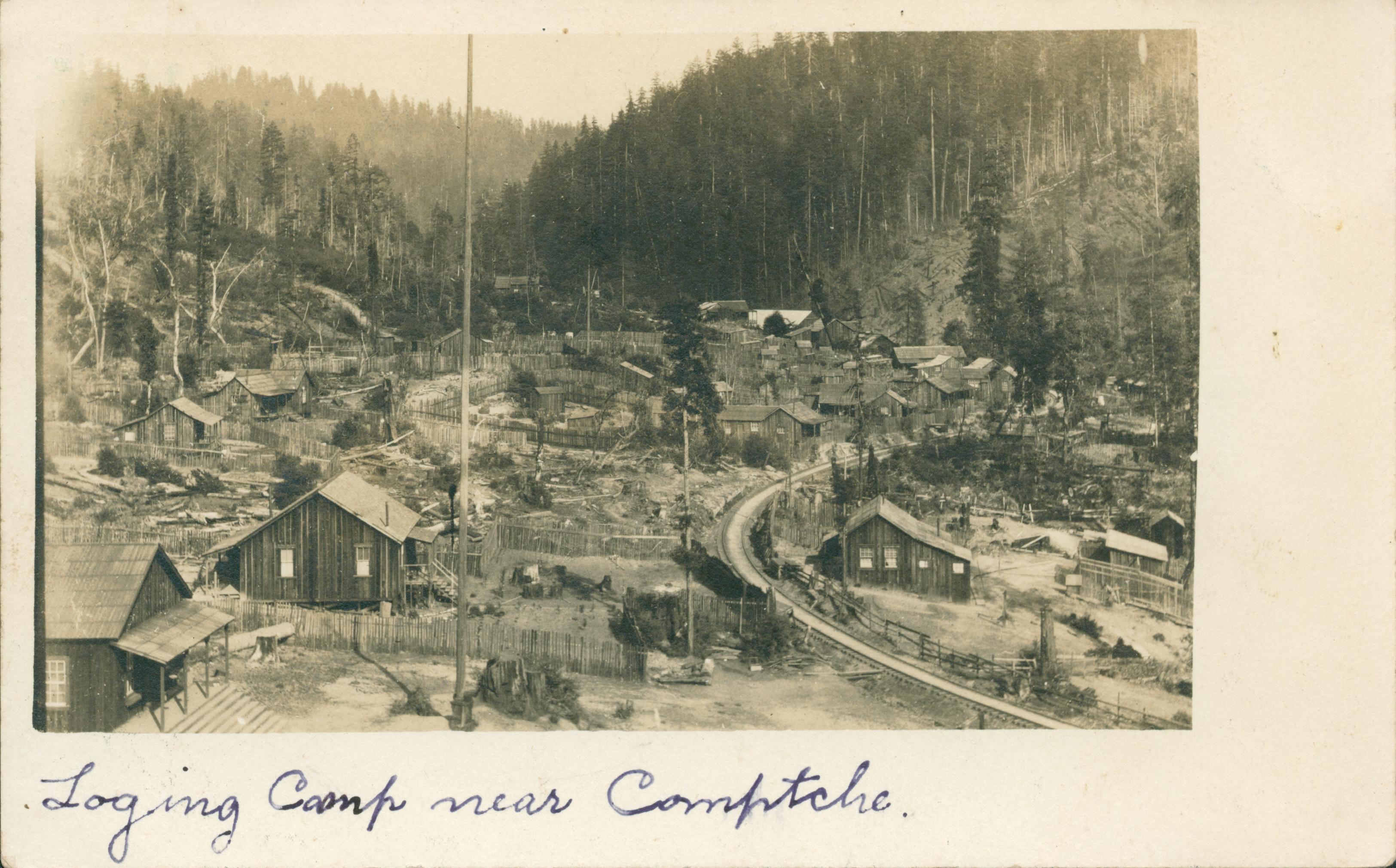 Logging camp in valley, surrounded by tree covered mountains, train track runs through camp