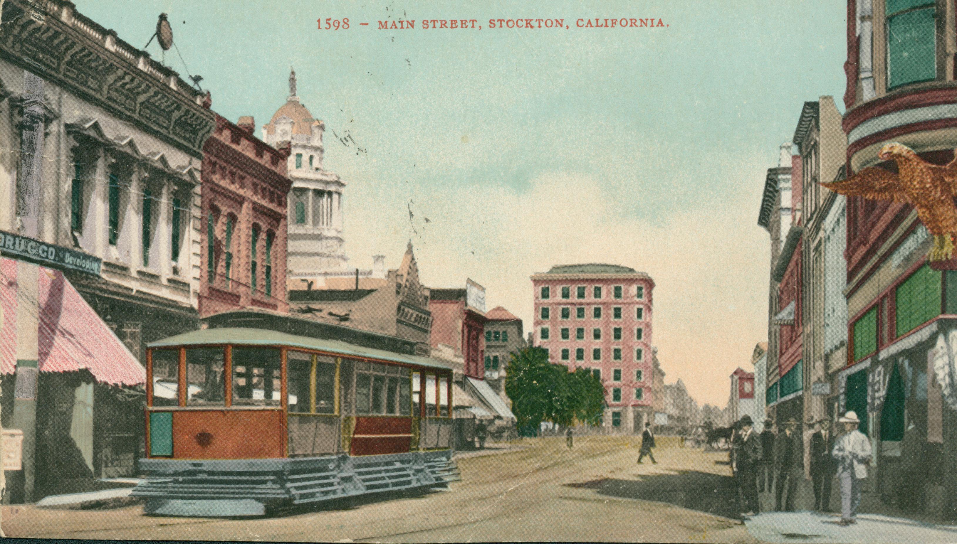 View of Main Street, buildings on either side of the street with a trolley and tracks down the center of the street, people waiting on the corner
