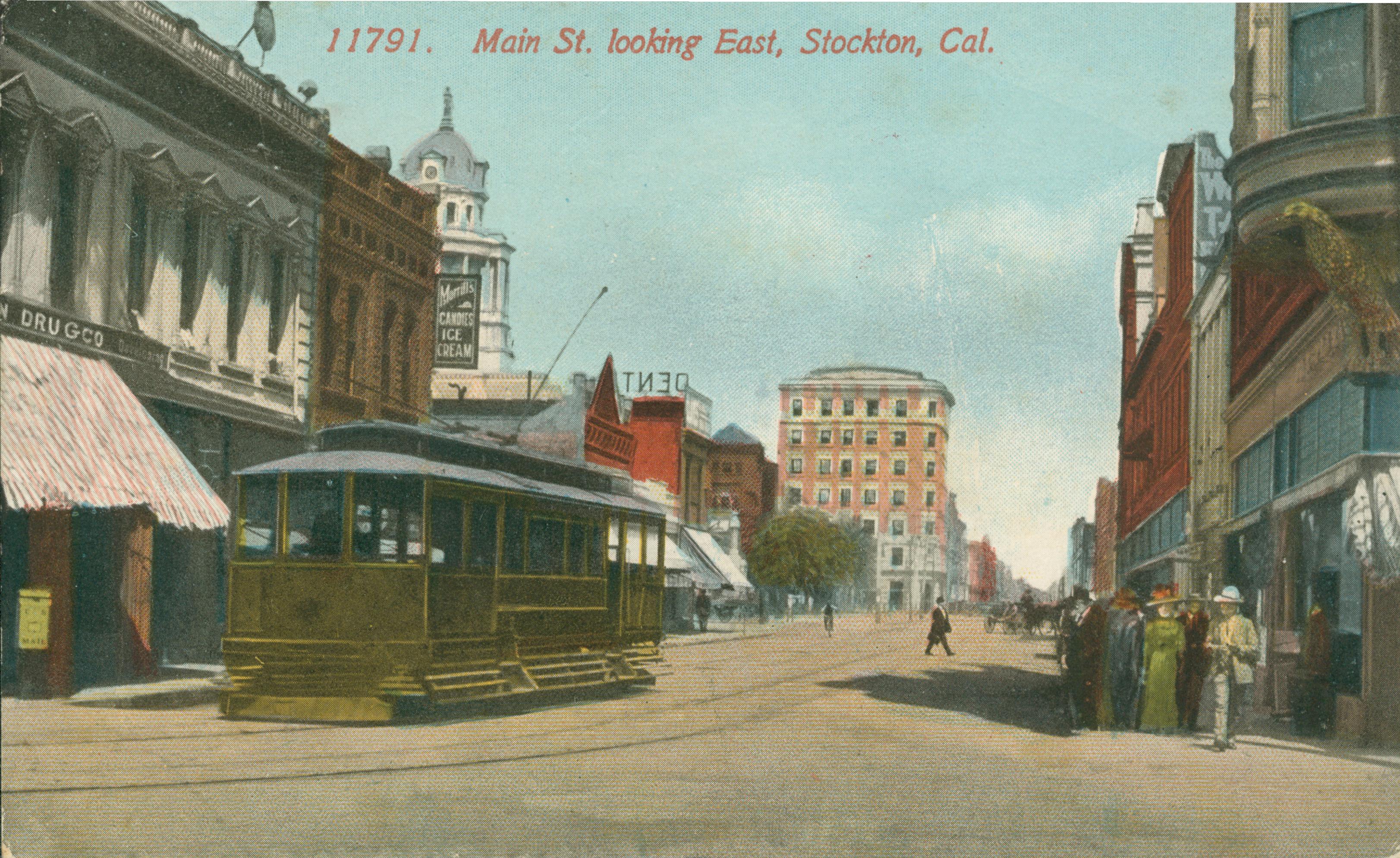 View of Main Street, buildings on either side of the street with a trolley and tracks down the center of the street, people waiting on the corner