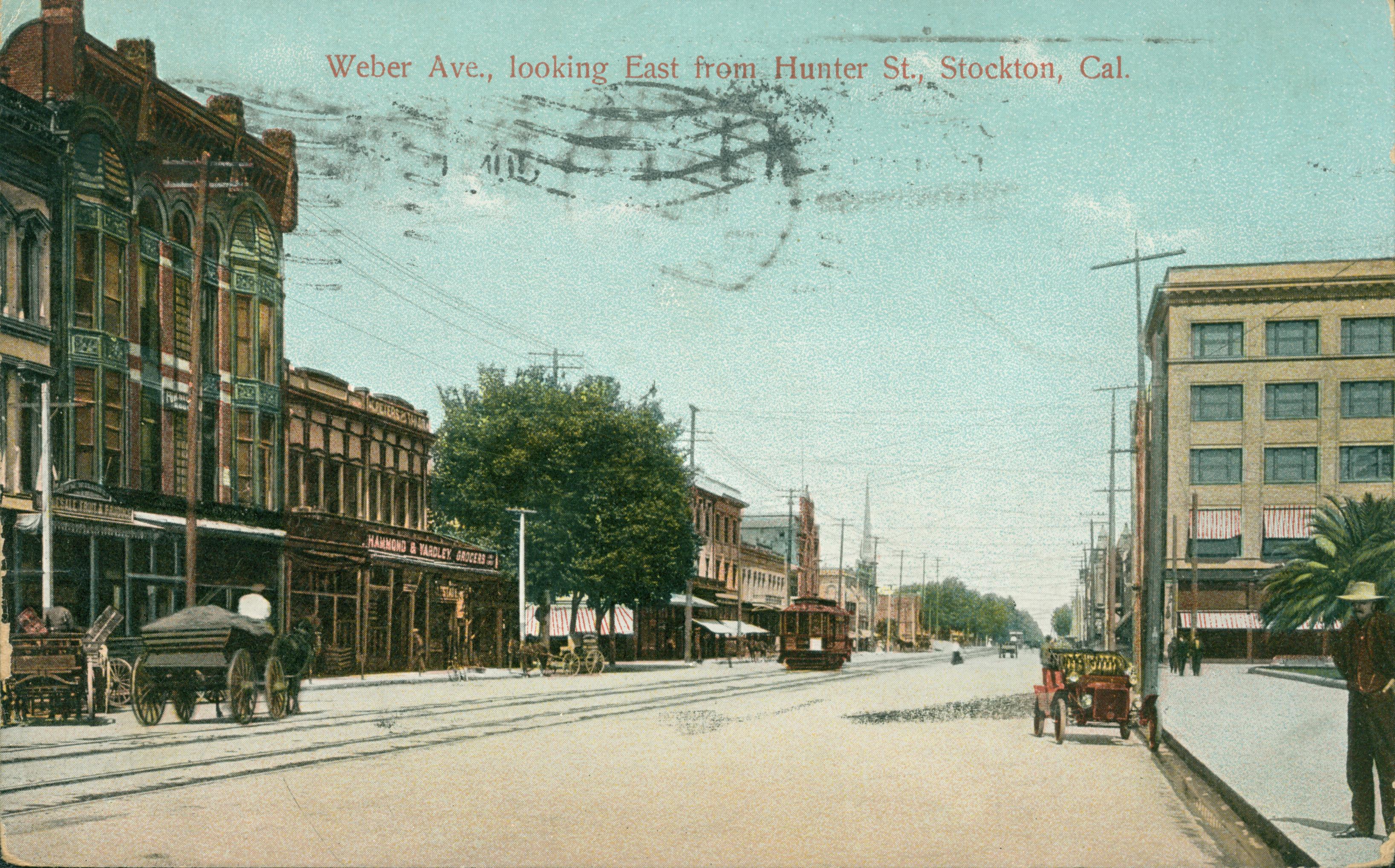 View of Main Street with trolley car and tracks down the center of the street, person waiting on corner, automobiles and wagons along side the street