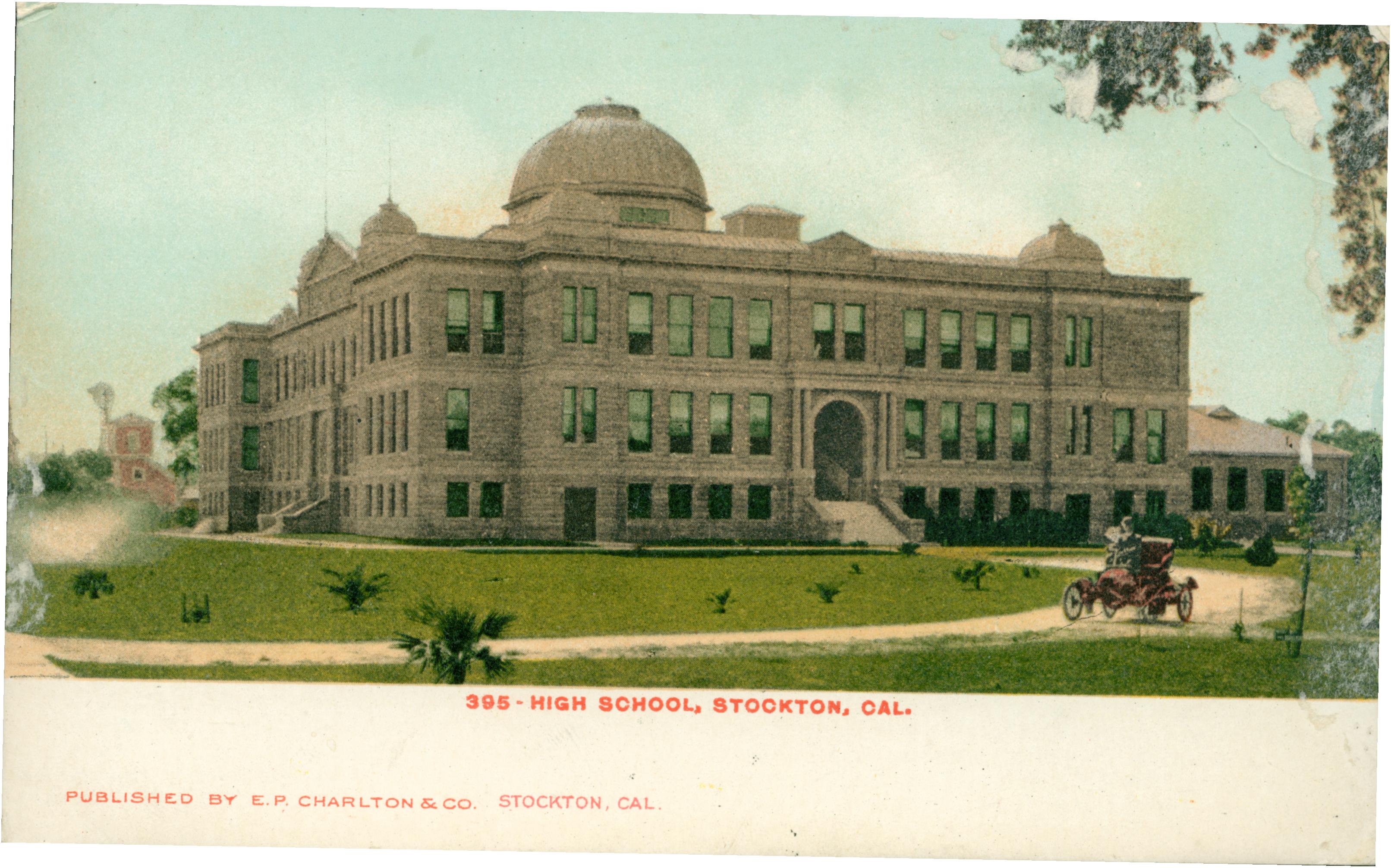 Exterior view of the high school, multi-story building, large grass lawn with small dirt drive, one lone automobile on the road