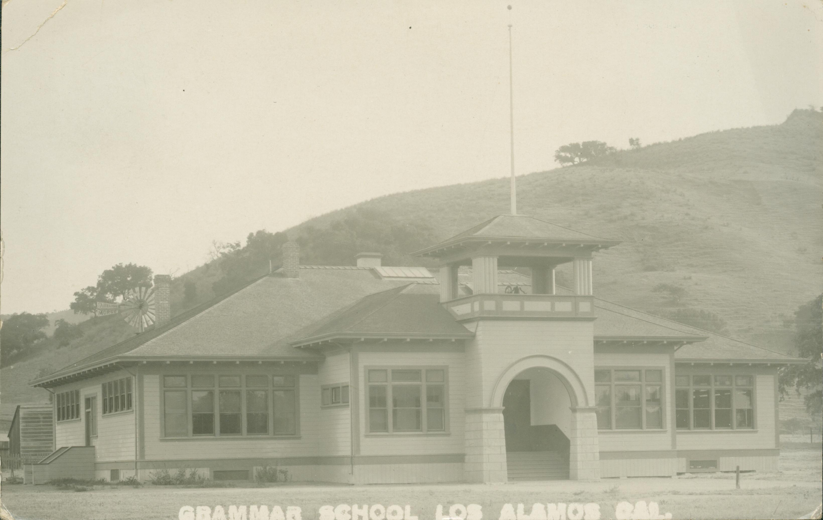 Exterior view of single story grammar school, windmill behind school, large hill with few trees behind school