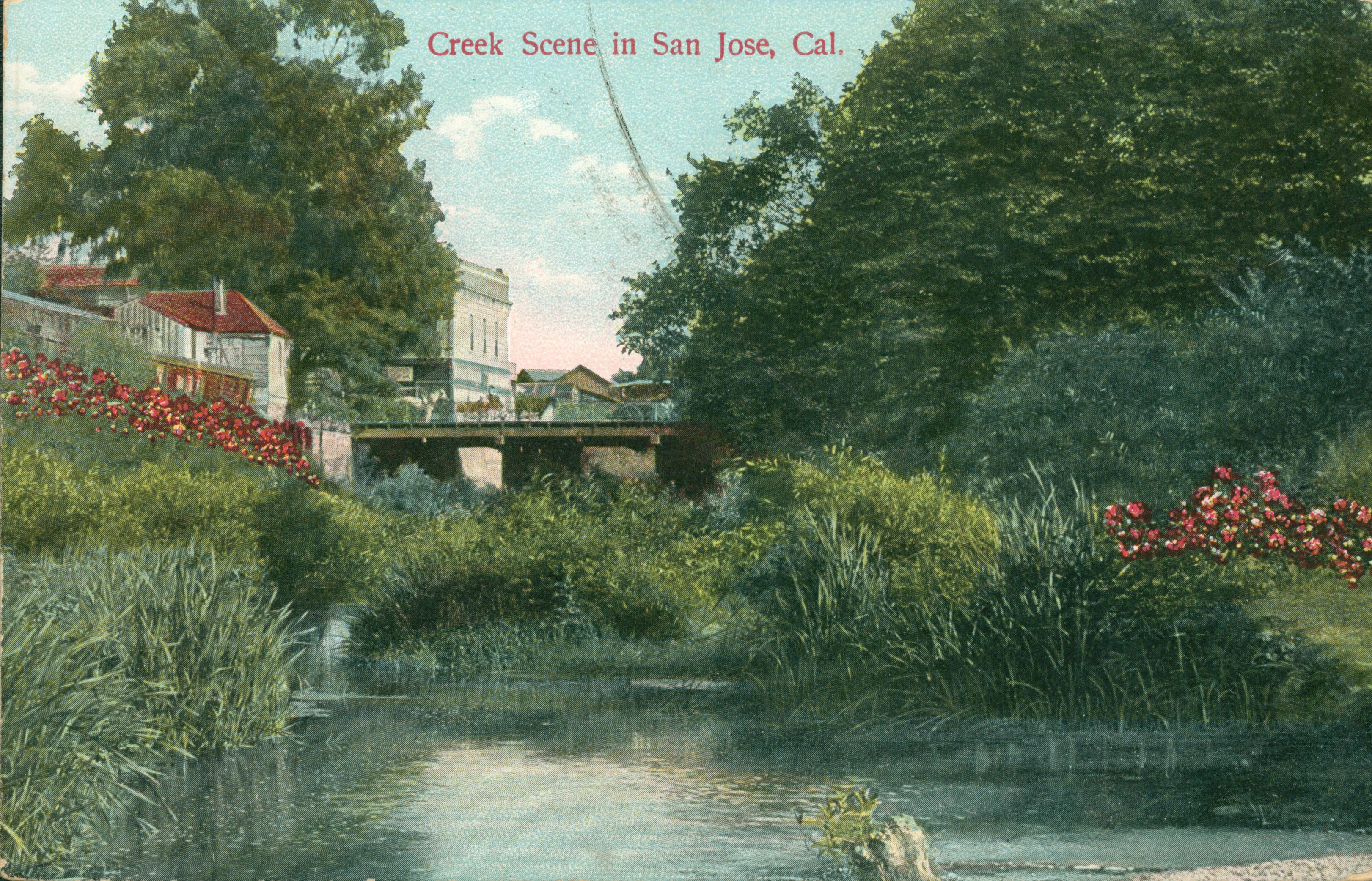 View of small creek surrounded by reeds, bushes and flowering bushes, buildings are high upon a bank with a bridge crossing the creek, horses lined up are on the bridge.  Large trees are in the background