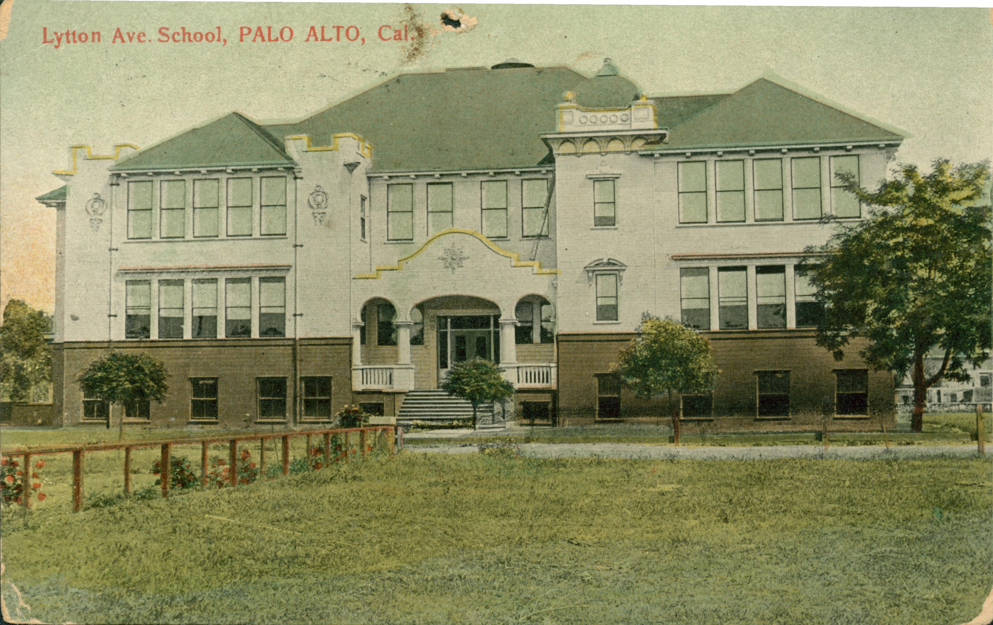Exterior view of Lytton Ave. School, large lawn and trees bordering the school, fence down the center of the lawn with flowering bushes, driveway separating grass area from building