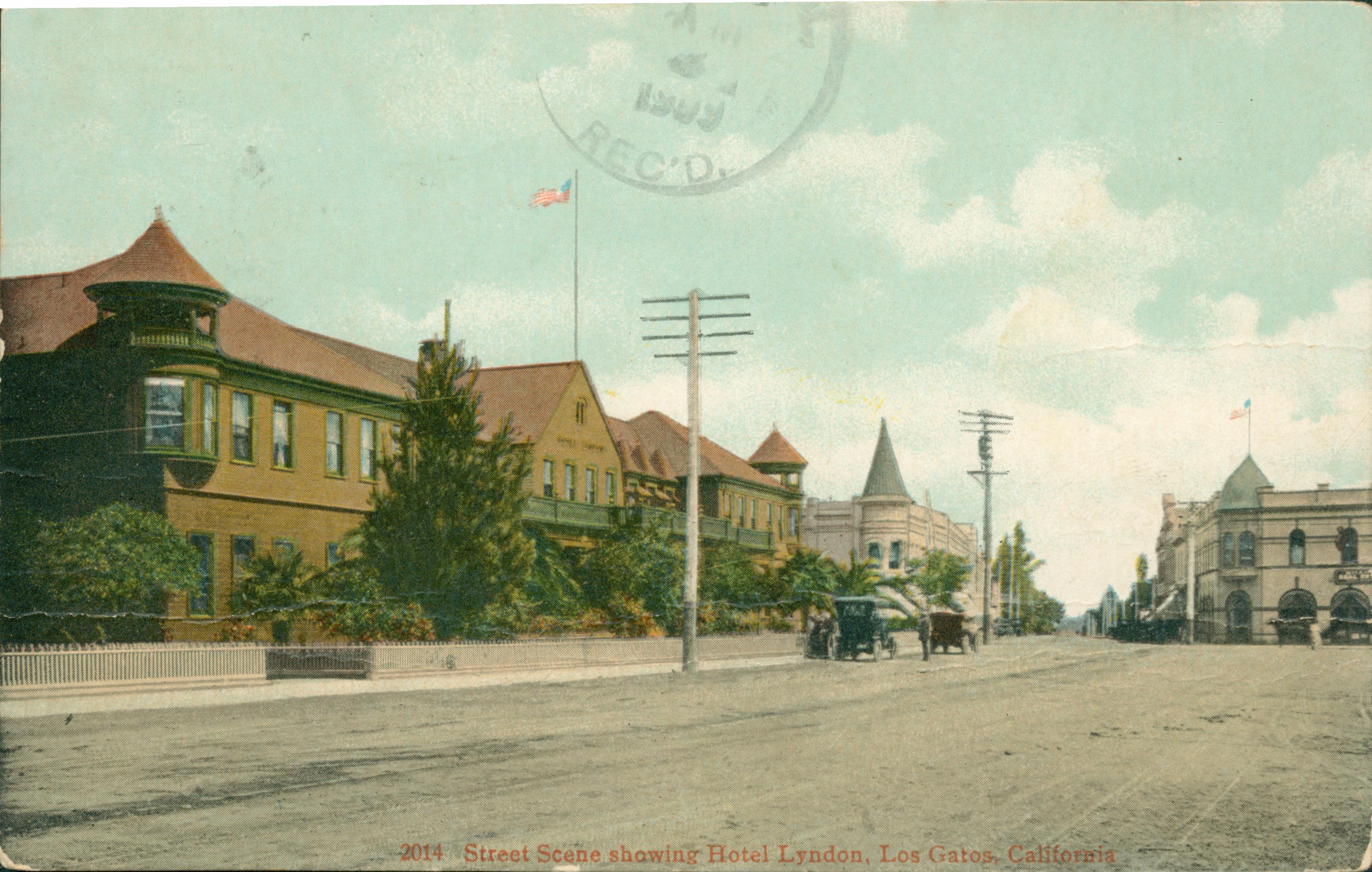 Large buildings with trees and picket fences, large dirt boulevard with automobiles parked on the side. Tall electric poles on side of road, flag flying from atop building