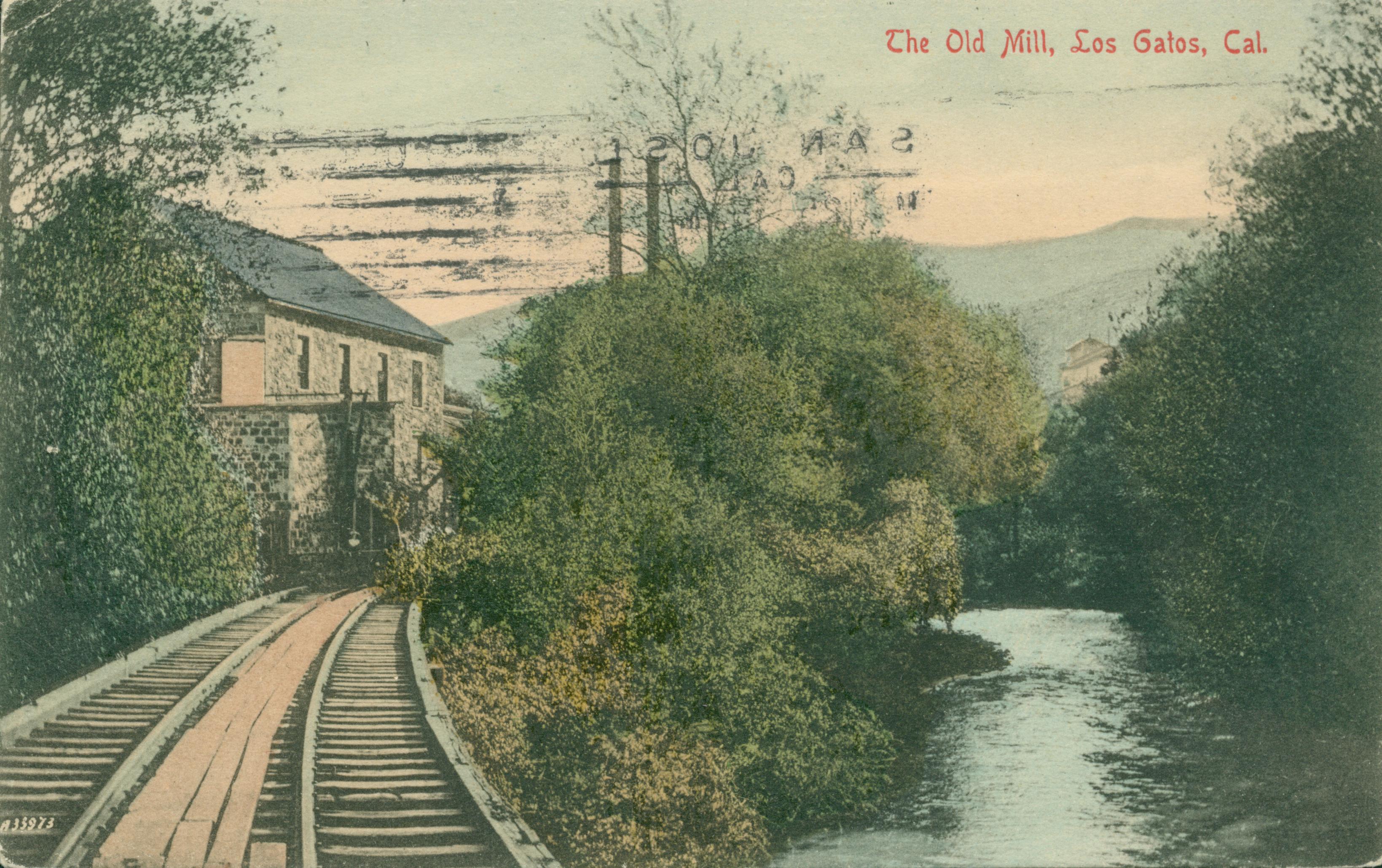 Double railroad tracks leading to stone mill, bushes and tree surrounding building and track, small creek to the right of the tracks and mill, trees on either bank of the creek, hills in the background