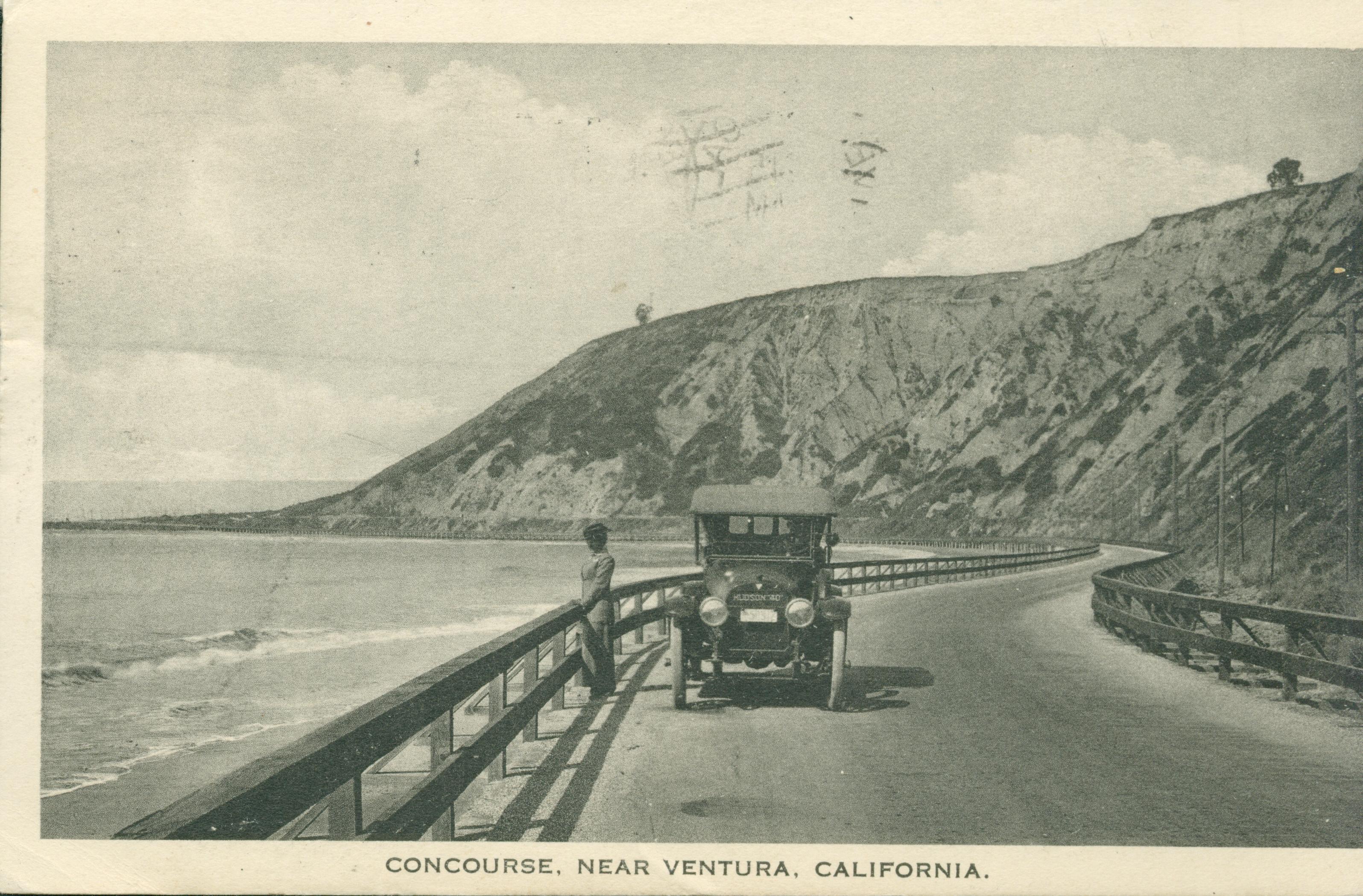 Shows a woman looking down at the beach by a car, stopped in the middle of a coastal highway