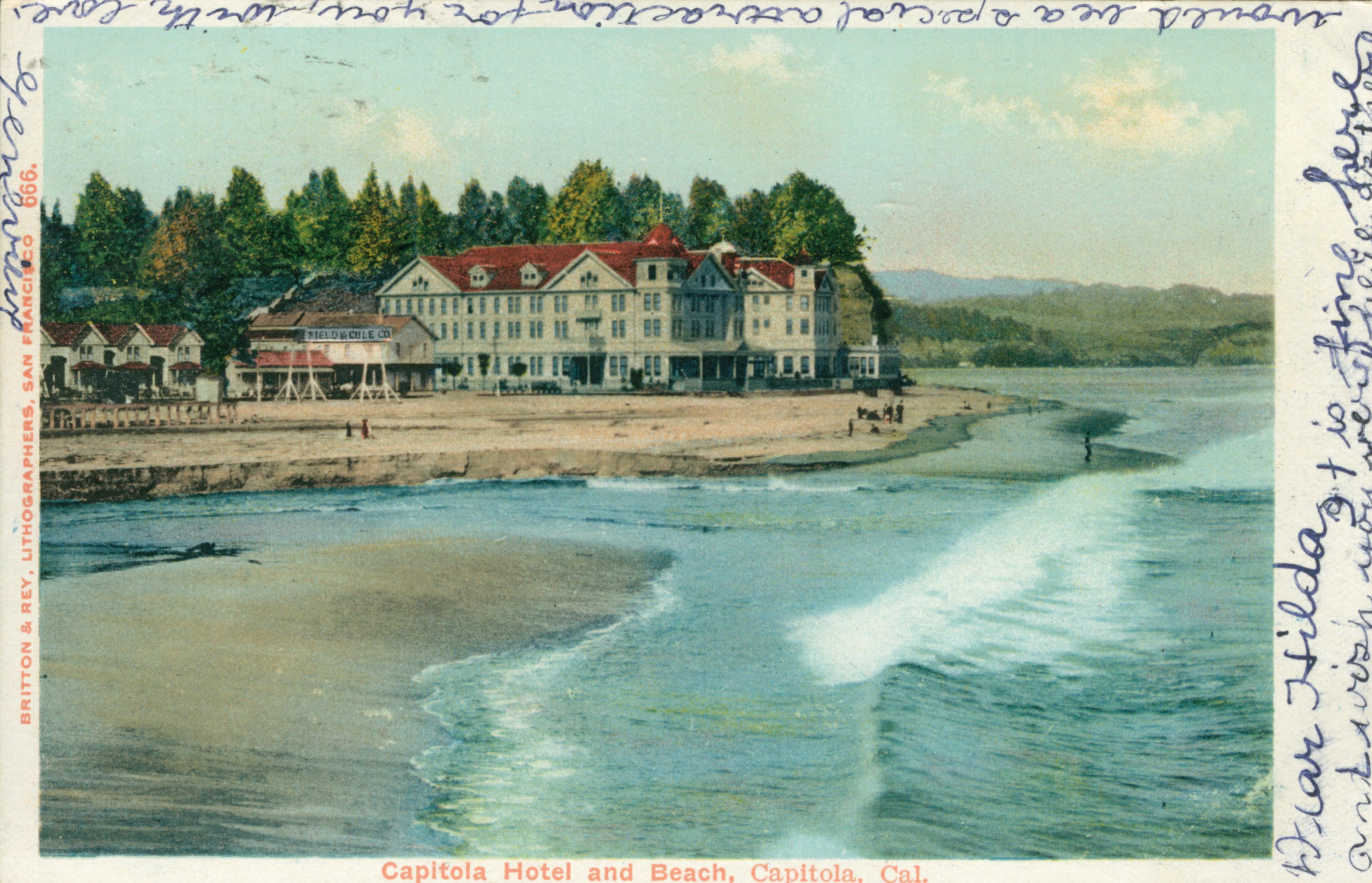 Capitola Hotel on the beach, people walking on the beach, river outlet leading to the river, trees in the background